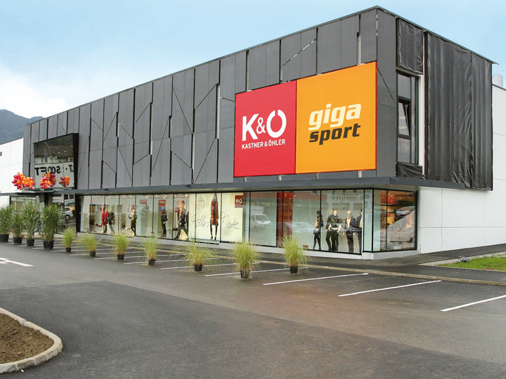 Un moderno edificio comercial con un gran letrero rojo anaranjado que dice "K&O Kastner & Öhler" y "giga sport". La fachada exterior tiene paneles oscuros y grandes escaparates con maniquíes. En primer plano se ve un pequeño aparcamiento.