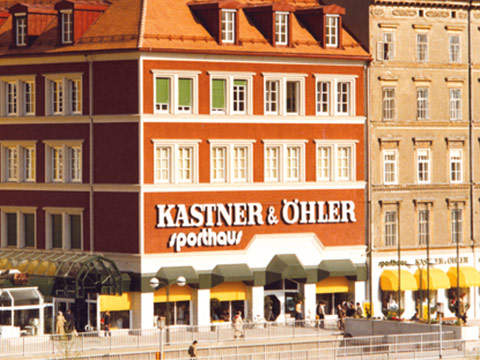 Ein großes Gebäude aus rotem Backstein mit zahlreichen Fenstern trägt an seiner Fassade „Kastner & Öhler / Sporthaus“. Im Untergeschoss befindet sich ein Ladenlokal mit gelben Markisen und Menschen, die in der Nähe in einer sonnigen Umgebung spazieren gehen.