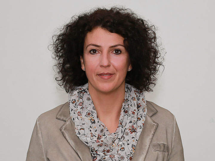 Una persona con cabello oscuro y rizado, que lleva un pañuelo floral y un blazer beige, mira directamente a la cámara. El fondo es simple y neutro.