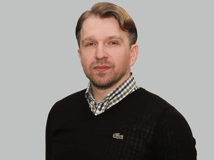 Un hombre con pelo corto y barba lleva un jersey negro sobre una camisa a cuadros. Está de pie frente a un fondo gris liso y mira a la cámara.