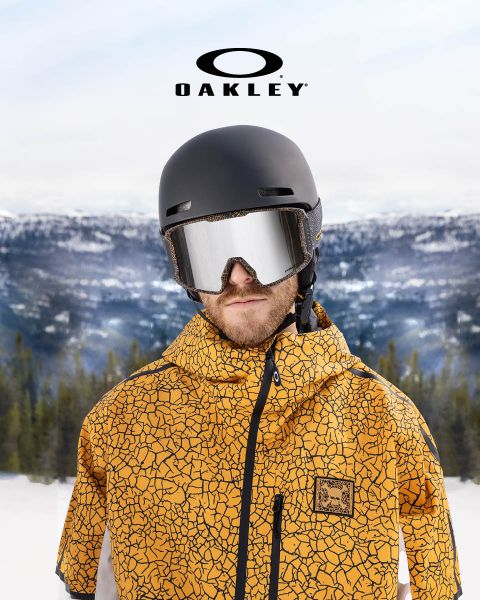 Una persona con un casco negro, gafas protectoras reflectantes y una chaqueta amarilla con un patrón crepitante está de pie frente a un paisaje montañoso nevado. En la parte superior de la imagen se ve el logotipo de Oakley.