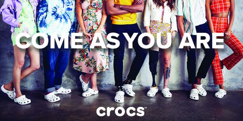 Un grupo diverso de personas está de pie junto a una pared de hormigón. Cada uno lleva un atuendo diferente y Crocs blancos. El texto "VEN COMO ERES" se ve claramente encima del logotipo de Crocs.