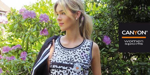 Una mujer con una blusa sin mangas con un patrón blanco y negro está de pie al aire libre junto a flores púrpuras florecientes y plantas verdes. En el lado derecho se ve el logo "CANYON women sports". Ella sostiene una chaqueta negra sobre su hombro.
