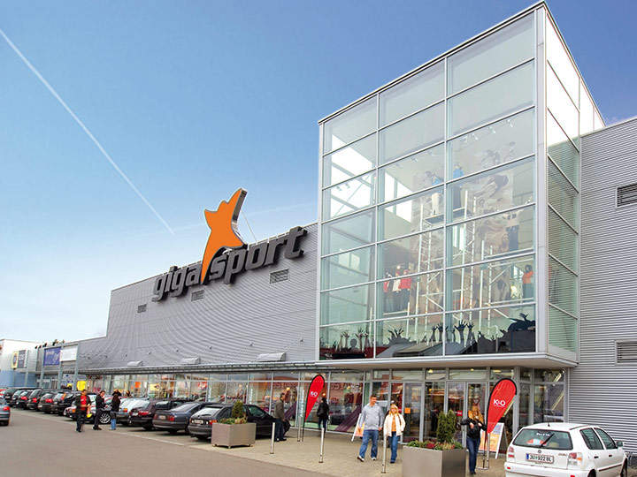 Vista exterior de una gran tienda de artículos deportivos con fachada de cristal y un logotipo naranja. Varios coches están aparcados en el estacionamiento y algunas personas se dirigen a la entrada. El edificio tiene un diseño moderno bajo un cielo azul despejado.