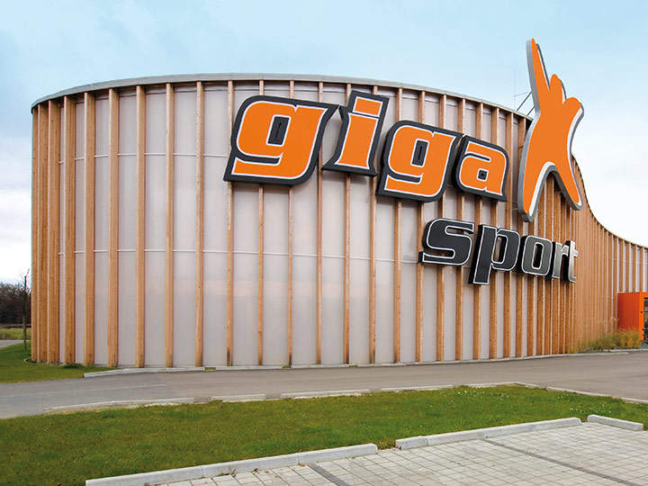 Vista exterior de un edificio grande y redondo con una fachada revestida de madera. Un gran letrero naranja y negro con la inscripción "Gigasport" cuelga del edificio. El entorno incluye un camino pavimentado y una zona de césped.