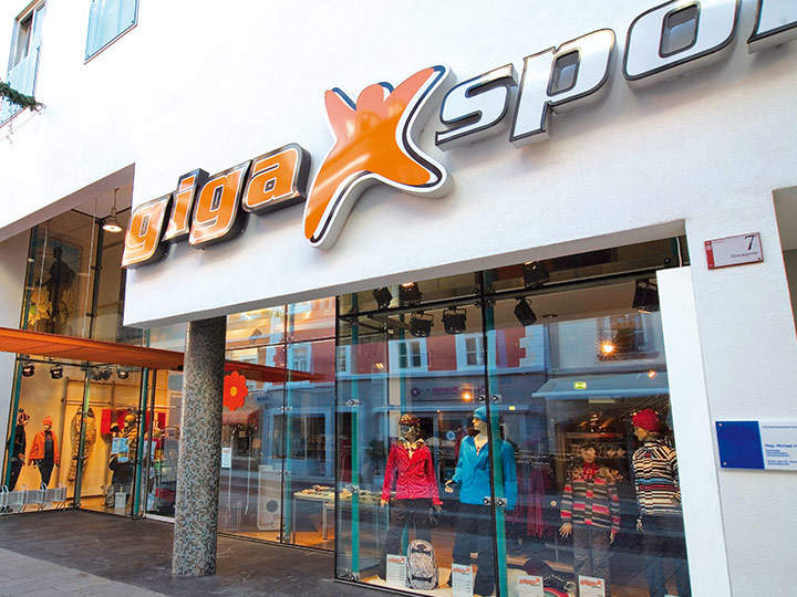 Vista de la calle de una tienda Gigasport con una fachada moderna. La entrada está flanqueada por grandes ventanales de cristal que muestran maniquíes vestidos con ropa deportiva colorida. Un letrero llamativo de "Gigasport" está colocado sobre la puerta.