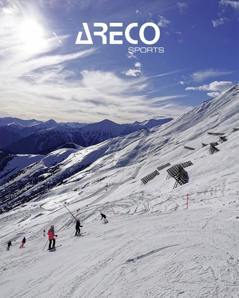 Esquiadores en una ladera nevada bajo un cielo azul con nubes dispersas. El sol brilla intensamente y proyecta sombras sobre la nieve. En la parte superior está el texto "ARECO SPORTS". En el fondo se ven picos nevados.