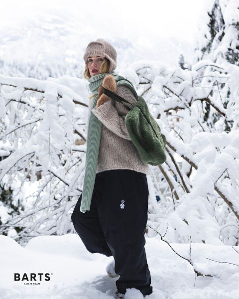 Una persona con un gorro de punto beige, suéter, bufanda verde y pantalones negros está de pie en un paisaje nevado y lleva una mochila verde. Ramas y árboles cubiertos de nieve forman el fondo. El texto en la imagen dice "BARTS AMSTERDAM".