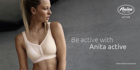 Mujer en sujetador deportivo de pie frente a un fondo gris mirando hacia un lado. El texto dice "Mantente activa con Anita Active". El logo de Anita y el enlace al sitio web están en la esquina.