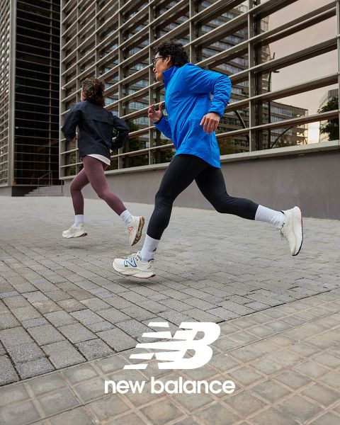 Dos personas trotan al aire libre, una con una chaqueta azul y leggings negros, la otra con una chaqueta negra y leggings color castaño. Ambas llevan zapatillas blancas. Corren junto a un edificio moderno con paneles de vidrio. Abajo se puede ver el logotipo de New Balance.