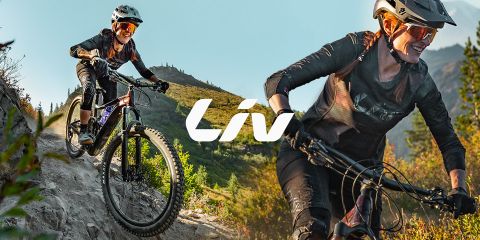 Dos mujeres montan en bicicleta de montaña por un sendero accidentado con árboles y montañas al fondo. Ambas llevan cascos y gafas de sol. En el centro se puede ver claramente la palabra "Liv".