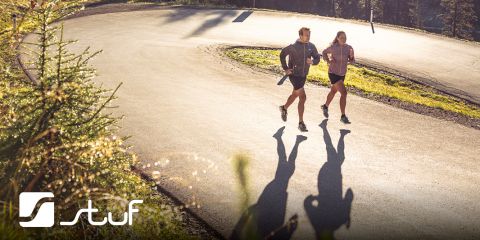 Dos personas trotan por una carretera sinuosa rodeada de un bosque. El sol proyecta largas sombras sobre la acera. Ambos visten ropa deportiva. En la esquina inferior izquierda hay un logotipo que dice "Stuf".
