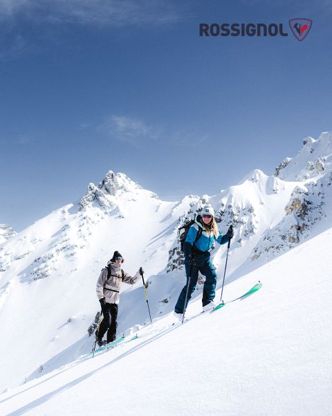 Dos esquiadores con ropa de invierno suben una ladera nevada en un día despejado. Al fondo se ven picos escarpados y nevados contra un cielo azul. El logo de Rossignol se ve en la esquina superior derecha.