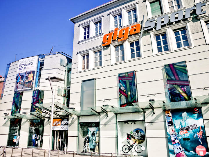 Vista de la calle de la tienda Gigasport con grandes ventanales que exhiben productos deportivos. Una valla publicitaria muestra a corredores. El edificio tiene una fachada moderna con "Gigasport" en letras llamativas de color naranja y gris.