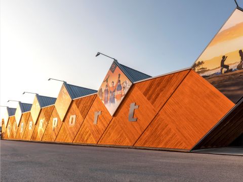 Edificio moderno con fachada hexagonal de madera y el letrero "ggysport". En la parte superior se ven grandes carteles, incluyendo imágenes de personas practicando actividades al aire libre. En el fondo se ve un cielo azul brillante.