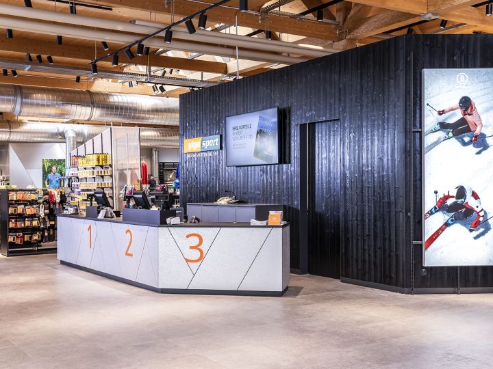 Un interior moderno de una tienda de deportes con un elegante mostrador de atención al cliente con las inscripciones 1, 2, 3. En la pared detrás se ven imágenes deportivas y hay estantes ordenados con varios artículos deportivos. El techo tiene vigas y tuberías a la vista.