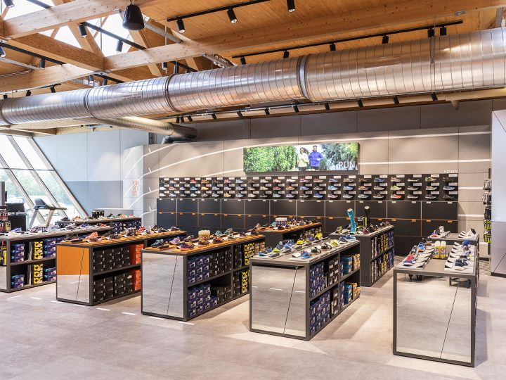 Una zapatería moderna con techos de madera, tuberías a la vista y grandes ventanales. Exhibidores de zapatos con varios estilos de calzado están distribuidos por todo el espacio. Una pantalla digital en la pared trasera muestra una imagen de personas al aire libre.