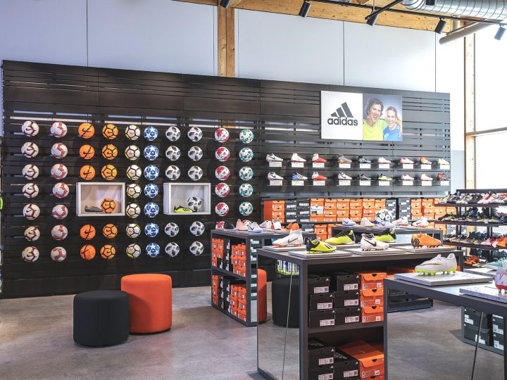Interior de una tienda de deportes con una exhibición en la pared de varios balones de fútbol y estantes con zapatos Adidas. Debajo, cajas de zapatos de color naranja brillante apiladas ordenadamente. La tienda tiene un diseño moderno con detalles en madera e iluminación industrial.