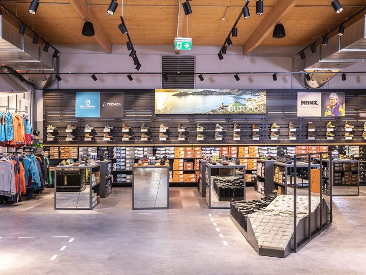 Una zapatería moderna con una selección de calzado de exterior en estanterías. Las marcas son visibles en un expositor de pared. La tienda cuenta con una iluminación elegante, accesorios de espejo y una zona para probarse los zapatos en una rampa texturizada. La ropa se encuentra en el lado izquierdo.