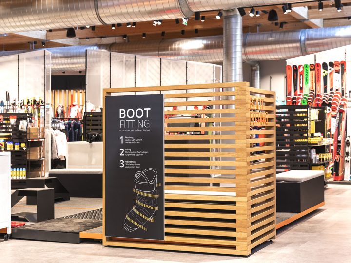 Un área de prueba de botas en una tienda de esquí con una elegante estructura de madera y un letrero negro. En el fondo, se ven varios equipos de esquí, incluyendo esquíes y bastones, ordenados en estantes.