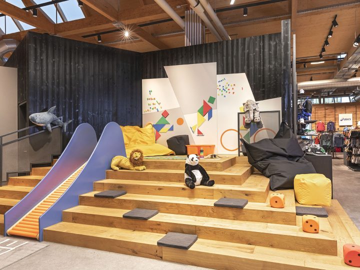 Parque infantil con escalones de madera, un tobogán azul y coloridos diseños geométricos en la pared. En los escalones hay peluches, incluyendo un león y un panda, así como pufs. El espacio está en un ambiente industrial moderno con tuberías a la vista y detalles en madera.