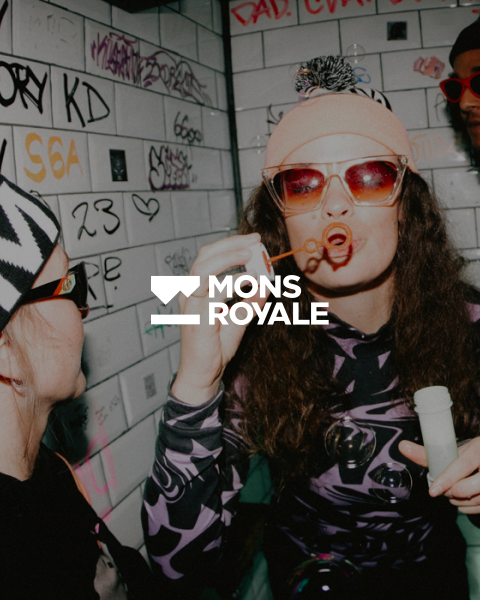 Dos personas en una habitación decorada con grafitis. Uno lleva gafas de sol y gorro y hace pompas de jabón, mientras que el otro con sombrero estampado y gafas de sol observa. En el centro está el texto "MONS ROYALE".