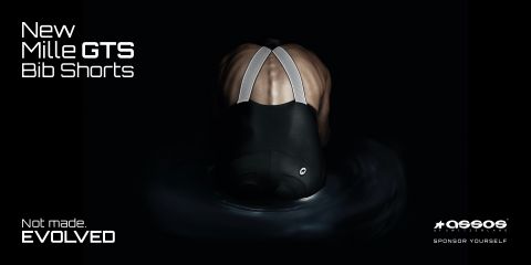 Se ve a una persona de espaldas con culotes de ciclismo, en la esquina superior izquierda está el texto "New Mille GTS Bib Shorts". La imagen tiene un fondo oscuro que resalta los tirantes reflectantes y el material del pantalón.
