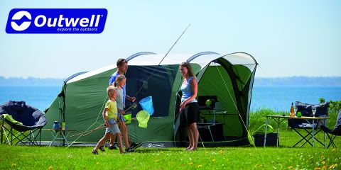 Una familia de cuatro miembros disfruta de un día soleado acampando junto al agua. Dos niños sostienen redes de pesca, mientras que dos adultos están de pie junto a ellos. Una gran tienda de campaña verde está montada, rodeada de sillas de camping y equipo. El logo de Outwell se ve en la esquina superior izquierda.