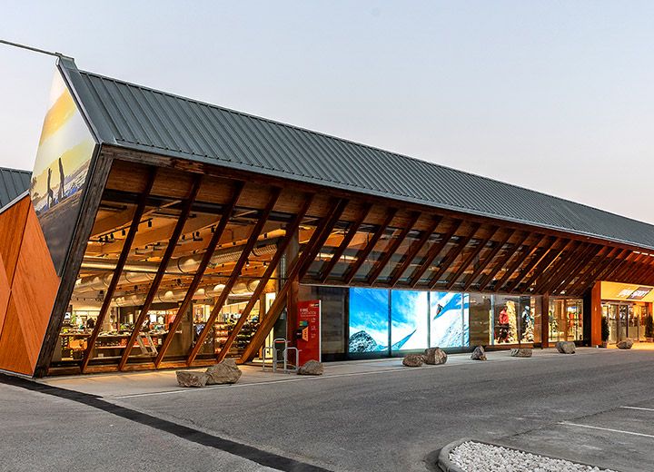 Un edificio moderno con una construcción de techo inclinado única con grandes ventanales y elementos de madera contra un cielo despejado. En la esquina superior derecha hay un letrero de "Ganador del Premio de Diseño Alemán 2020". El exterior está cálidamente iluminado.