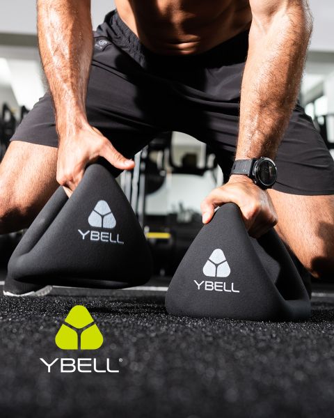 Una persona en ropa de entrenamiento sostiene dos pesas YBELL negras en el suelo del gimnasio. Lleva un reloj y pantalones cortos. El logotipo de YBELL se muestra en las pesas y en la esquina inferior izquierda.