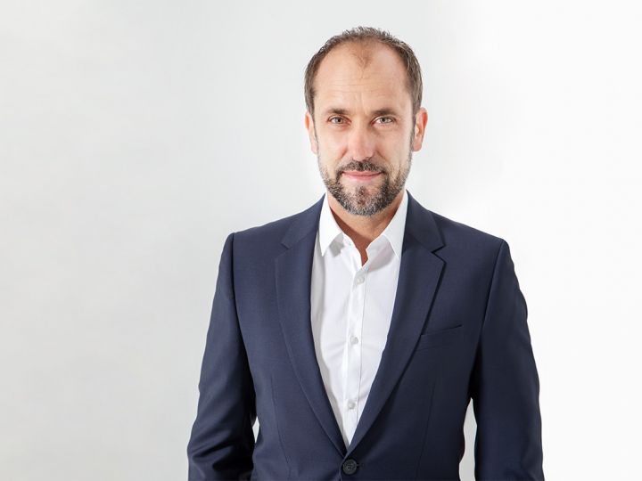 Un hombre con pelo corto y barba, vestido con un traje azul marino y una camisa blanca, está de pie frente a un fondo blanco liso y mira a la cámara con una leve sonrisa.