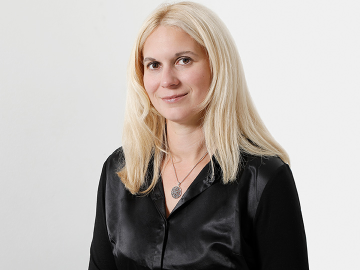 Una persona con cabello largo y rubio, que lleva una blusa negra y un collar con un colgante redondo, está de pie frente a un fondo blanco sencillo y sonríe levemente.