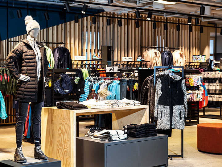 Vista interior de una tienda de ropa con un maniquí que lleva una chaqueta negra, un sombrero blanco y botas. Varias chaquetas coloridas y ropa deportiva se exhiben en estantes y perchas, con detalles de madera e iluminación moderna al fondo.