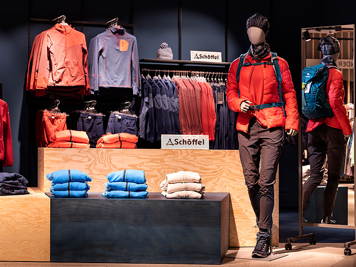 Maniquí con ropa de exterior con chaqueta naranja, pantalones negros, mochila azul y gorro de punto. En los estantes de exhibición hay prendas azules y beige dobladas y perchas con chaquetas. En el fondo se ve el logotipo de Schöffel.