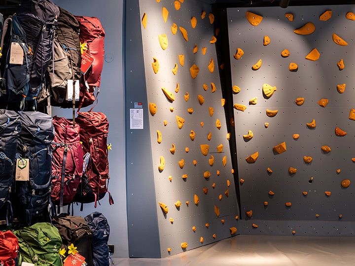 Pared de escalada interior con varias presas naranjas sobre un fondo gris. A la izquierda, varias mochilas de diferentes colores se exhiben en estantes. La iluminación es brillante y el suelo está pulido.