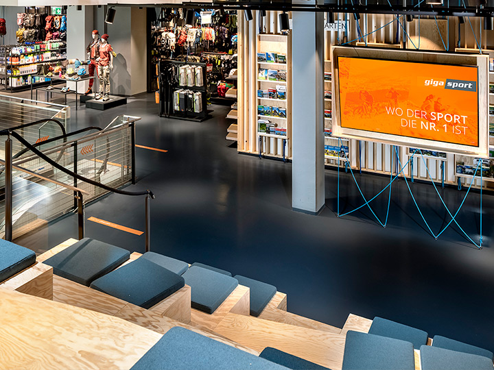 Vista interior de una tienda de deportes con asientos de madera con cojines, una gran pantalla con los anuncios "Gigasport" y "Donde el deporte es el número 1", escaleras mecánicas y artículos deportivos en exhibición. El espacio es moderno y bien iluminado.