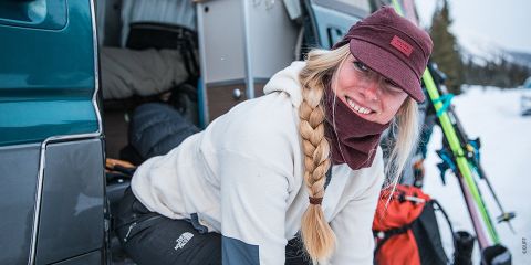 Una persona con una trenza larga y un gorro marrón castaño sonríe mientras sale de una camioneta. Está vestida con ropa de abrigo, rodeada de nieve, y se ven esquíes y equipo de nieve afuera.