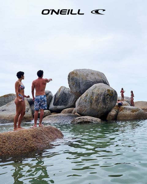 Una mujer con cabello largo está de pie en un paisaje desértico bajo un cielo azul con nubes dispersas. Lleva una camiseta sin mangas negra de O'Neill y se inclina ligeramente hacia adelante. En el lado derecho de la imagen se puede ver el logo de O'Neill.