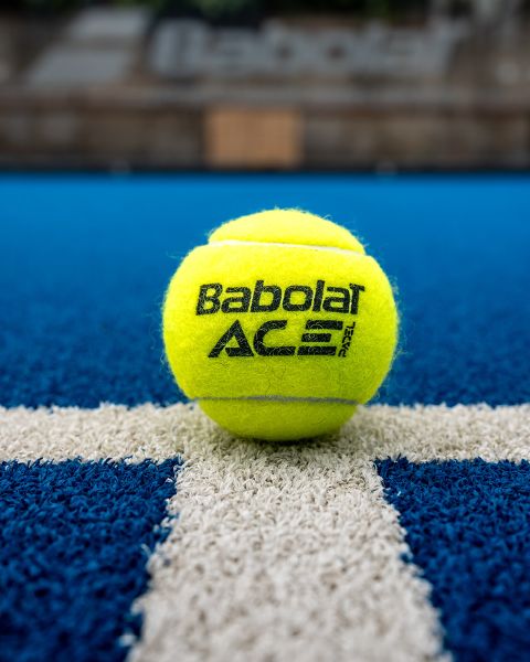 Una pelota de tenis Babolat amarilla está en una cancha de tenis azul, centrada en una línea blanca. El fondo está borroso, enfocando el logo de la marca y la textura de la pelota.