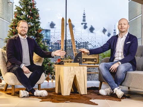 Zwei Männer sitzen in einem Innenraum mit einem Weihnachtsbaum auf der linken Seite. Sie lächeln und zeigen auf einen Tisch mit einer Holzauszeichnung und einem Retro-Radio. Große Fenster geben den Blick auf eine verschneite Stadtlandschaft im Hintergrund frei.