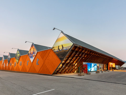 Einzigartiges modernes Gebäude mit schrägem Dach und dreieckigen Abschnitten, mit großen Wandgemälden von Landschaften und Silhouetten im Freien. Das Gebäude hat markante Fenster und einen klaren Himmel im Hintergrund, mit einem Parkplatz im Vordergrund.