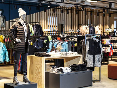 In einem Bekleidungsgeschäft ist eine Schaufensterpuppe mit einer schwarzen Pufferjacke, Strickmütze, Leggings und Stiefeln zu sehen. Auf dem Holztisch liegen gefaltete Kleidungsstücke, während auf den Regalen im Hintergrund verschiedene Jacken und Oberteile ausgestellt sind.