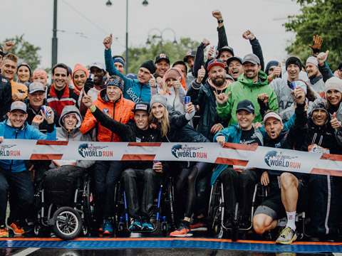 Eine bunt gemischte Gruppe von Menschen posiert aufgeregt an der Startlinie eines Laufevents. Einige sitzen im Rollstuhl. Sie tragen Sportkleidung und halten große Energydrink-Dosen in den Händen. Vor der Gruppe ist ein Banner mit der Aufschrift „World Run“ zu sehen.
