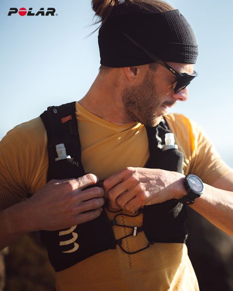 Una persona con una cinta negra en la frente y gafas de sol se ajusta su chaleco de hidratación negro. Lleva una camisa amarilla y un reloj inteligente en la muñeca. El logo de Polar se ve en la parte superior izquierda. La luz del sol ilumina la escena.