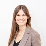Mujer sonriente con cabello castaño largo, lleva un blazer a cuadros y una blusa negra, y está de pie frente a un fondo blanco liso.