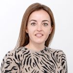 Una mujer con cabello castaño sonríe a la cámara. Lleva un jersey beige con un patrón de rayas de cebra negras. El fondo es blanco liso.