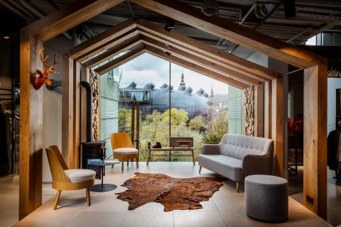 Modernes Interieur mit Holzbalken in Hausform, mit grauem Sofa, beigen Stühlen, rundem Ottoman und Tierfellteppich. Der Blick aus dem Fenster zeigt üppige Bäume und ein einzigartiges Gebäude mit einem Turm im Hintergrund. Ein orangefarbener Hirschkopf schmückt die Wand.