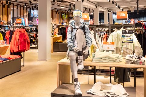Innenansicht eines Sportartikelladens mit einer Schaufensterpuppe in Outdoor-Ausrüstung, darunter Windjacke, Shorts und Beanie. Sie sitzt auf einem Ausstellungstisch mit gefalteter Kleidung und verschiedenen Kleidungsstücken im Hintergrund.