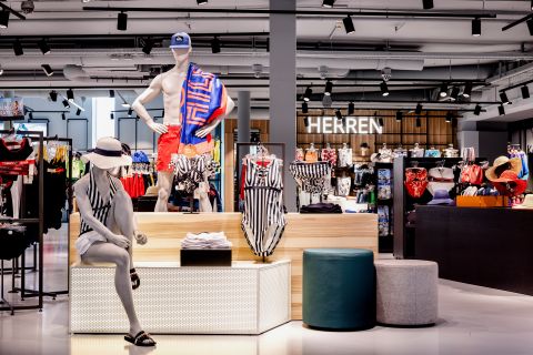 In einem Schaufenster eines Einzelhandelsgeschäfts sind Schaufensterpuppen in Strandkleidung zu sehen, darunter ein gestreifter Badeanzug und ein gemusterter Schal. Im Hintergrund sind Ständer mit bunter Kleidung unter dem Schild „HERREN“ zu sehen. Der Boden ist glänzend und in der Nähe gibt es ein paar runde Sitze.