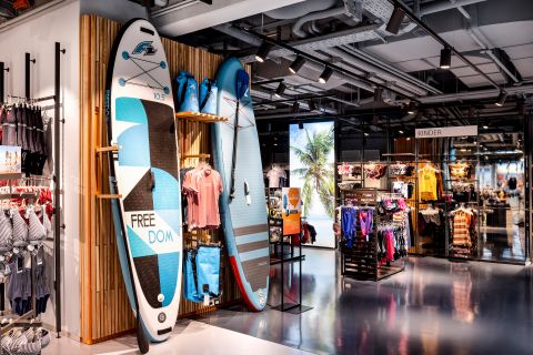 Innenansicht des Ladens mit zwei Stehpaddelbrettern, die an einer Holzwand lehnen. In der Nähe stehen Kleiderständer mit Kleidung in verschiedenen Farben. Über der Kinderbekleidung ist ein „Kinder“-Schild zu sehen und eine Auslage mit Strandmotiven sorgt für tropische Stimmung.